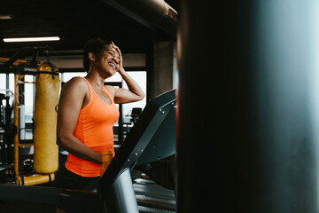 Fototapeta na wymiar Satisfied fit woman standing on treadmill in the fitness gym and feeling exhausted after running. She is wiping the sweat from her forehead and smiling.