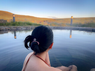 Young woman sitting in an outdoor hot spring In the background is a blurred view of mountains and fields.