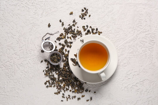 Cup of tea with infuser and dry green tea leaves on white textured background