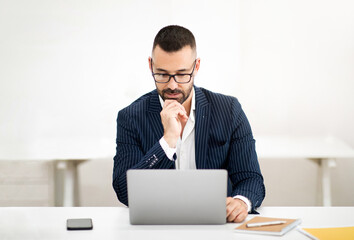 Pensive serious caucasian mature man in suit, glasses at table looks at laptop, have video call