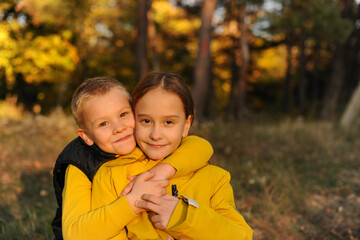 Little  brother and his  sister are hugging against the backdrop of the autumn forest. children are happy. The girl is wearing a yellow jacket the boy is wearing a black vest.