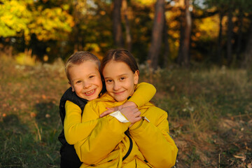 Little  brother and his  sister are hugging against the backdrop of the autumn forest. children are happy. The girl is wearing a yellow jacket the boy is wearing a black vest.