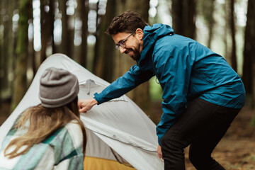 Cheerful young caucasian couple in jackets in forest enjoy travel vacation, make tent