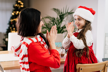 Happy mother and daughter in Santa hat playing clapping hands game and have fun on decorated modern open space kitchen while drinking cocoa. Family holidays. Festive christmas mood. Selective focus.
