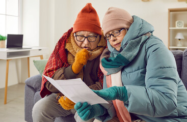 Portrait of a senior couple man and woman sitting in the living room at home in winter outerwear and looking at the utility bill warming up wrapped in a blanket. Heating problems concept.