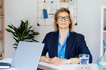 Smiling mature businesswoman with laptop at her office workplace, confident middle-aged experienced...
