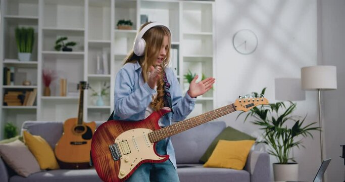 Energetic funny girl with long hair playing electric guitar, sing song and dancing at living room at home. Attractive girl rocker of 11-12 years old playing rock music on electric guitar. Slow motion