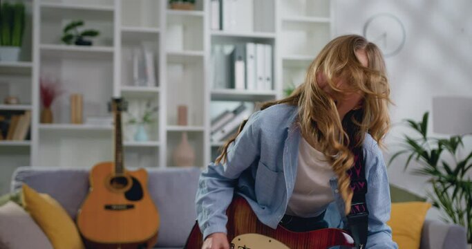 Close up of long haired young woman rocker in denim shirt playing acoustic guitar, singing and dancing at light living room at home