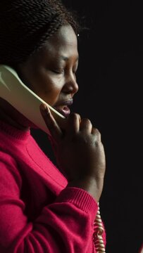 vertical video Side-view portrait of black woman with pink sweater standing with handset wired phone unsatisfied by the talk. Young african american female having a serious conversation on landline