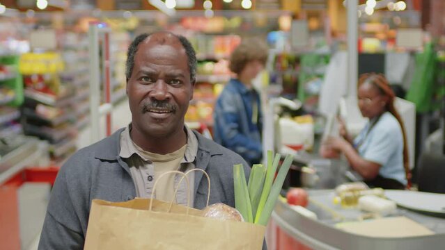 Selective focus portrait of senior Black man holding paper bag full of food products standing in supermarket looking at camera