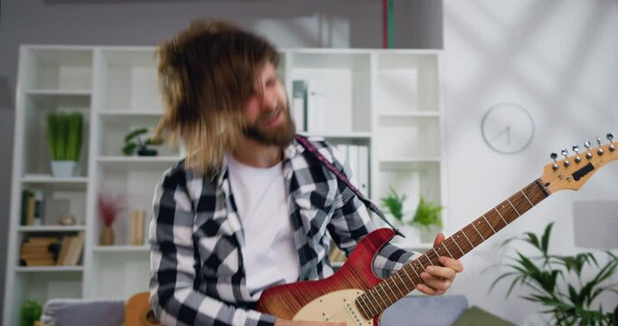 Excited young man in checkered shirt with long hair playing on electric guitar dancing and shaking head over in music studio, lifestyle