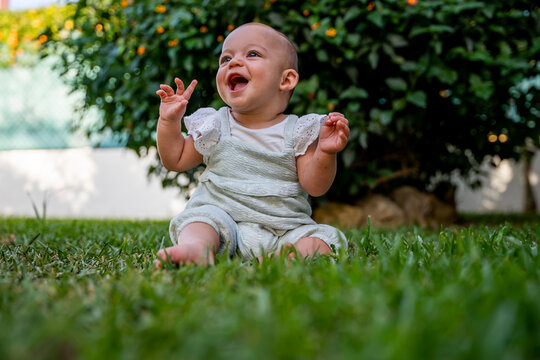 little baby sitting on the grass in the garden at home laughs out loud with her mouth open while raising her hands. concept fun in childhood