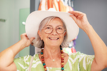 Cheerful senior woman in retail shop trying a white hat looking for sales and offers. Consumerism concept
