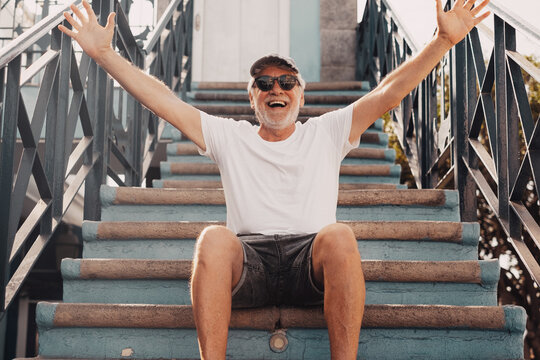 Happy senior handsome man with cap and sunglasses sitting with open arms in outdoors staircase. Carefree elderly man enjoying free time, vacation or retirement