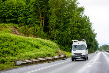 Compact cargo mini van with tubes on the roof running to side of the service driving on the wet highway road at rainy weather