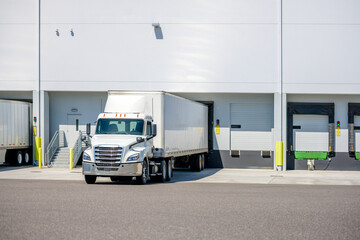 Day cab white big rig semi truck with dry van semi trailer standing on the warehouse dock gate loading cargo for the next commercial delivery