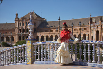 Obraz premium Young black and South American woman in a beige gypsy flamenco suit and red shawl, dancing with a fan in a beautiful square in the city of Seville in Spain. Concept dance, folklore, flamenco, art.