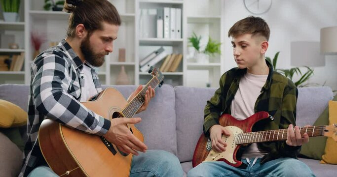Talented music teacher and teenage student boy sitting on sofa learning to play on guitar in private lesson in light living room or music studio. Slow motion