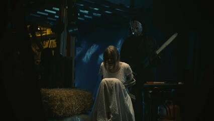 Female tied with a rope on a chair in warehouse, man in the mask with a chainsaw standing behind her.