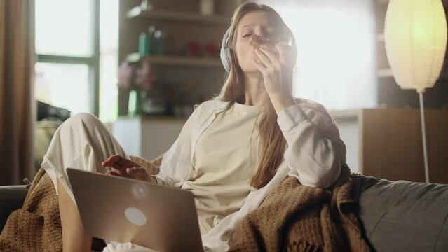 Pretty happy young woman freelancer with headphones listening music while working online typing browsing on laptop and eating pizza slice fast food enjoying great day at cozy home