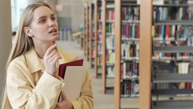 Smart pretty focused blonde girl student holding books looking away, walking in modern university campus library or bookstore thinking of college course study reading literature for doing research.