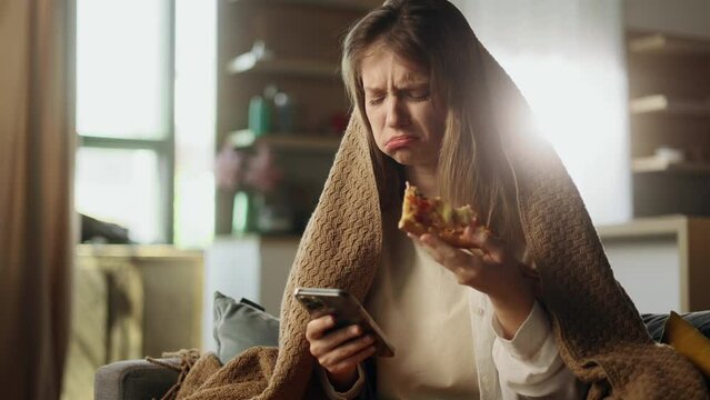 Portrait of sad crying young woman wrapped in blanket dealing with stress by eating food pizza and reading bad news email break up message from ex-boyfriend or personal problems at home