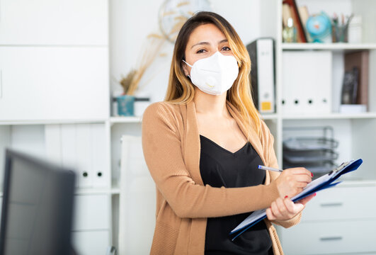 Young Latina Woman Secretary Wearing Face Mask Standing In Office With Clipboard Writing Instructions At Workplace