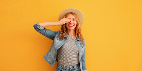 Portrait of beautiful happy surprised smiling young woman covering her eyes wearing summer straw hat, denim jacket on orange studio background