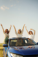 Obraz premium Happy young female friends in casual clothes and sunglasses raising arms in excitement while standing through window and roof of car while on vacation in countryside