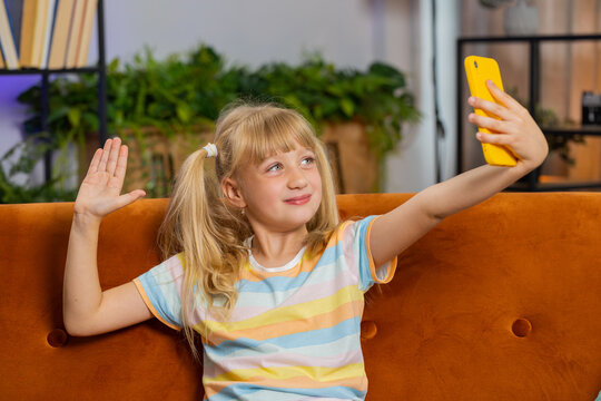 Young blonde child kid girl blogger taking selfie on smartphone, communicating video call online with social media subscribers. Female teen toddler recording video story at home in room sits on sofa