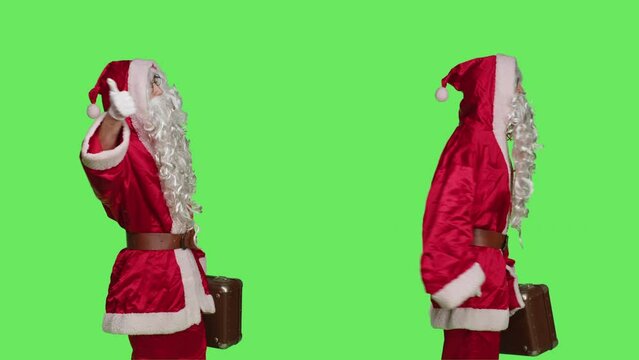 Santa claus with briefcase hitchhiking over greenscreen backdrop, asking to go somewhere while he is wearing festive costume with hat and beard. Confident man in cosplay suit with suitcase.