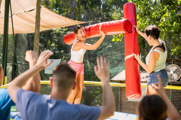 Fototapeta na wymiar Playful young woman having funny wrestling by inflatable logs with female friend in outdoor amusement playground on sunny summer day..