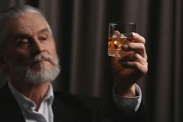 Senior man in suit holding glass of whiskey with ice cubes on brown background, selective focus
