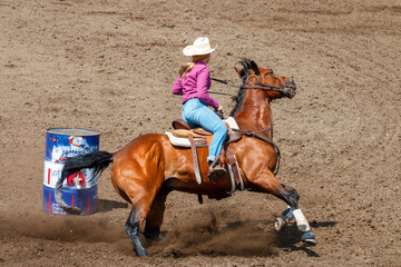 Cowgirl riding in a barrel racing completion at a rodeo. She is wearing red shirt and blue jeans and a white hat. The horse is brown. The barrel is behind the horse.