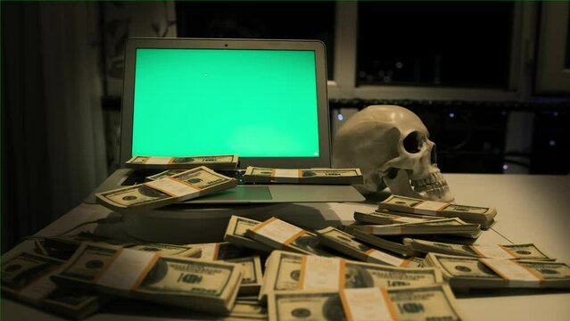 Laptop with green screen and new 100 dollars cash bills on desk next human skull in office night time. Internet fraud and bankruptcy. 
