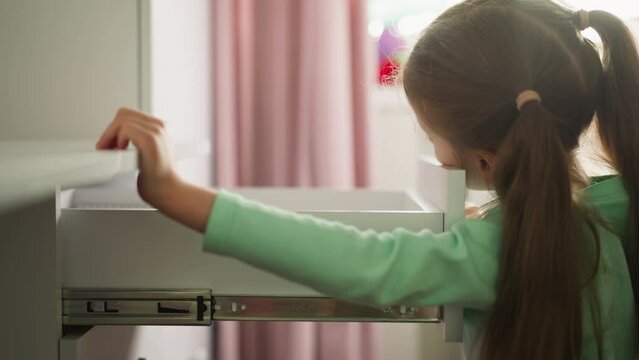 Curious little girl opens desk drawer in children room. Child seeks interesting items in commode container at home. Schoolgirl and domestic furniture