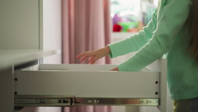 Little girl takes out files and papers of desk drawer in children room closeup. Child seeks supplies for schooling in commode box at home. Modern furniture