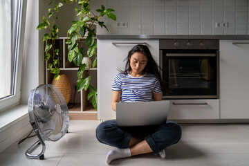Focused asian girl student with laptop is languishing heat sitting on cool floor next fan and open...