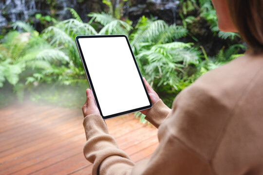 Mockup image of a woman holding digital tablet with blank white desktop screen in the garden with waterfall