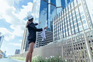 Expert female architect reviews Skytrain site, cross referencing blueprint for project approval.