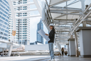 Expert female architect reviews Skytrain site, cross referencing blueprint for project approval.