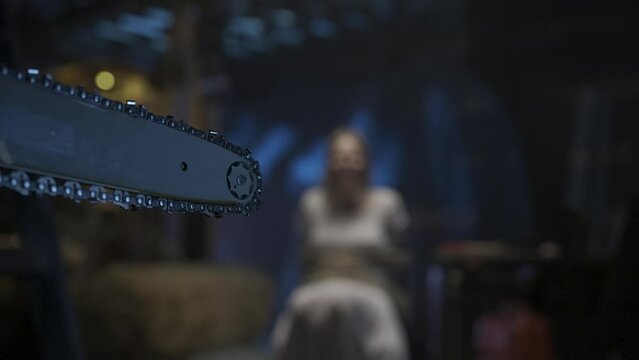 Closeup shot, focus on the working chainsaw. Girl victim tied with a rope sitting on a chair crying in screaming. HDR BT2020 HLG Material.