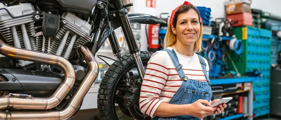 Photo sur Plexiglas Moto Portrait of smiling mechanic woman looking at camera while holding phone in her hands sitting over platform with custom motorcycle on factory