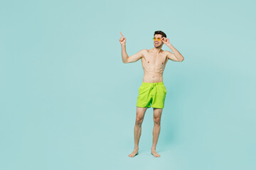 Obraz premium Full body young man wear green shorts swimsuit sunglasses relax near hotel pool look aside on area mockup point finger aside isolated on plain blue background Summer vacation sea rest sun tan concept