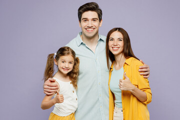 Young cheerful happy parents mom dad with child kid daughter girl 6 years old wear blue yellow casual clothes looking camera hug show thumb up isolated on plain purple background. Family day concept.