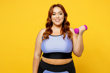Young happy chubby overweight plus size big fat fit woman wear blue top warm up training hold in hand dumbbell look camera isolated on plain yellow background studio home gym. Workout sport concept.