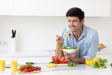Smiling man at kitchen home cooking tastes salad with fork, healthy eating