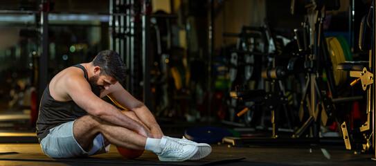 Caucasian man is facing overtraining syndrome after workout inside gym with dark background for...