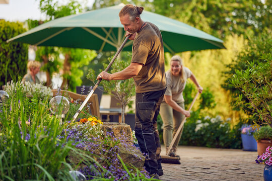 Male gardener raking the the flower bed while the collegue is sweeping the terrace in summer