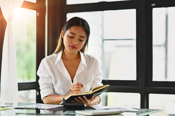 Obraz premium Millennial female employee checking work schedule plan on daily planner while sitting at modern workplace.